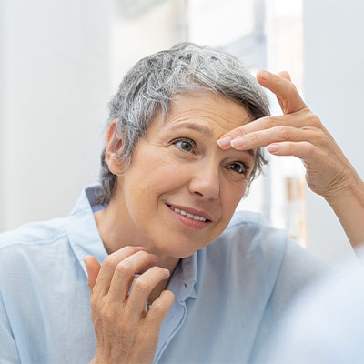 A woman with short gray hair looking into the mirror as she applies a Pevonia product.